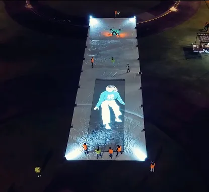 Vista aérea nocturna de una pantalla gigante extendida en el suelo, con un dron y personal alrededor
